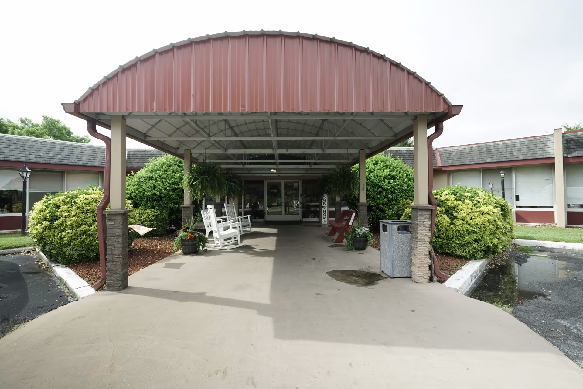 Entrance to Jefferson City Health & Rehabilitation Center featuring a covered drop-off area with a red metal roof, white rocking chairs, potted plants, and bushes on either side. The building has large windows and a concrete driveway leading up to the entrance.