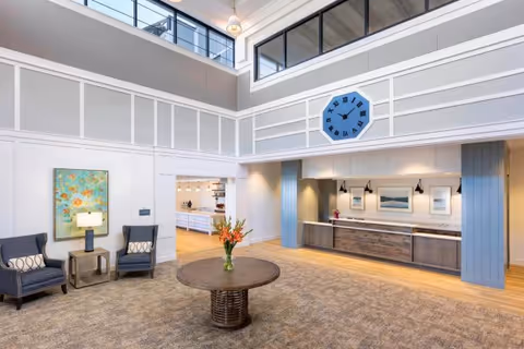 A spacious and well-lit interior common area with high ceilings and large windows near the ceiling. The room features a round wooden table with a vase of flowers in the center, two upholstered chairs with a small side table and lamp between them, a large blue clock mounted on the wall above a reception or service counter with framed artwork and pendant lights. The flooring is carpeted with a wood floor section near the counter and an open doorway leading to a kitchen or dining area.