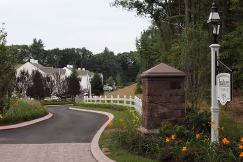 Curved driveway leading into Peachtree Village, bordered by a white fence and landscaped with orange flowers and greenery. A stone pillar and a lamp post with a Peachtree Village sign are visible on the right side, with trees and residential buildings in the background.