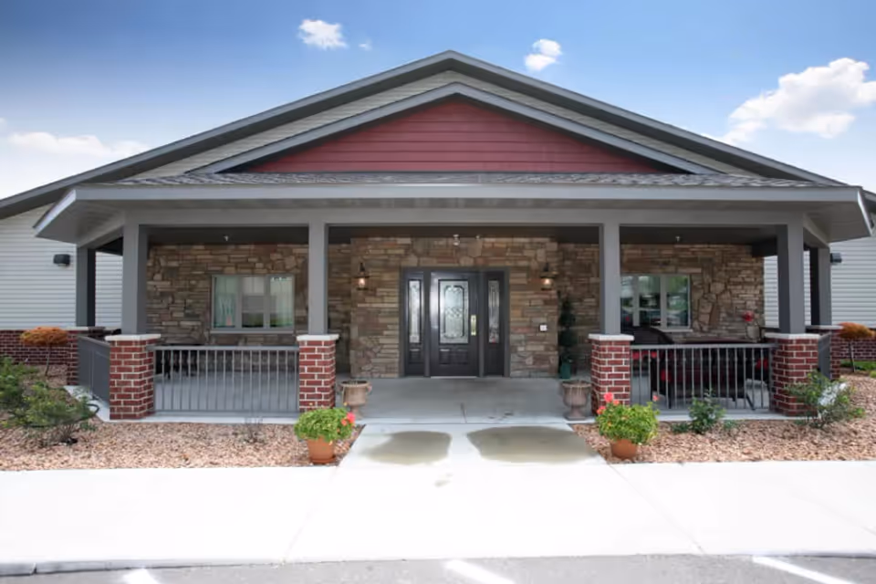 Front entrance of a single-story building with a covered porch, double doors, stone facade and potted plants.