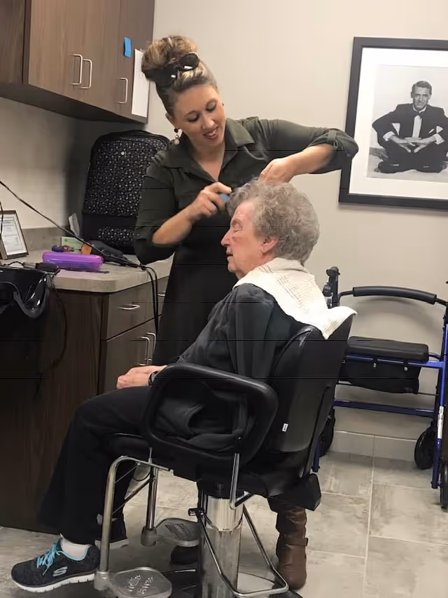 A woman is styling the hair of an elderly woman seated in a salon chair inside a room with cabinets and a framed black-and-white photo on the wall. A walker is visible in the background.