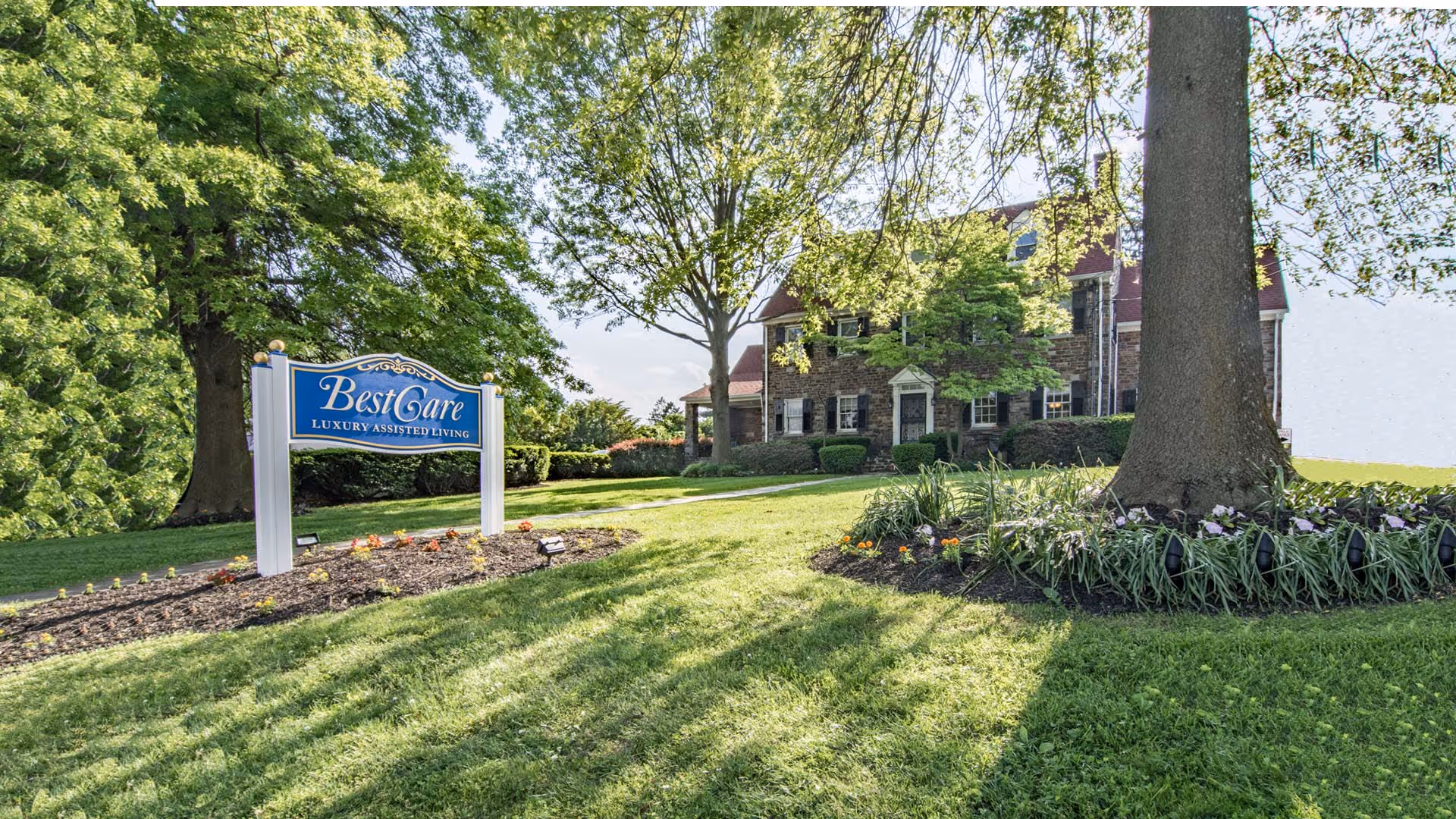 A large stone building with multiple windows and a red roof surrounded by green trees and well-maintained lawns. In the foreground, there is a blue and white sign that reads 'BestCare Luxury Assisted Living' placed in a flower bed with small plants and mulch.