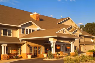 Exterior view of a senior living facility building with a large covered entrance, beige siding, stone accents, and a sloped roof under a clear blue sky.