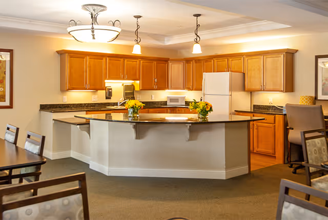 A spacious kitchen area with wooden cabinets, a white refrigerator, microwave, and a large island countertop with two vases of yellow flowers. The kitchen is well-lit with ceiling lights and pendant lights above the island. Surrounding the kitchen are dining chairs and tables.