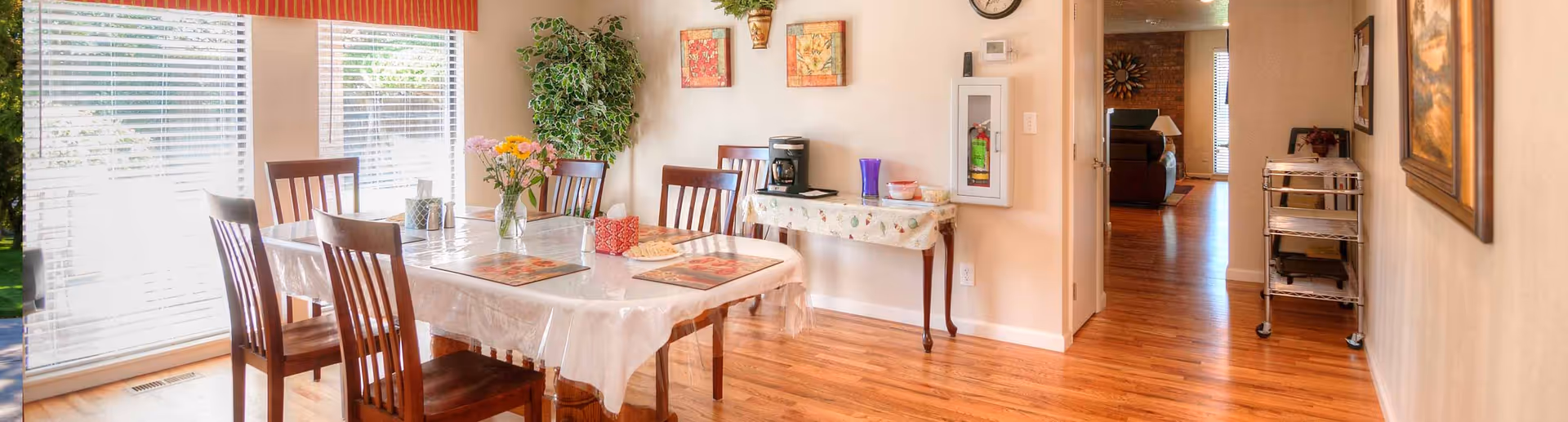 A bright dining room with a wooden table covered by a clear plastic tablecloth and four wooden chairs. The table is set with placemats, a vase of flowers, tissue boxes, and some snacks. Large windows with white blinds let in natural light. There is a small table against the wall with a coffee maker and some containers. The room has wooden flooring, a potted plant, and framed artwork on the walls. A hallway leads to another room with a clock on the wall and a metal cart on wheels.