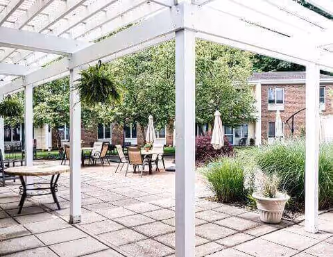 Outdoor patio area with a white pergola overhead, several tables and chairs arranged on a tiled floor, potted plants, and greenery including bushes and trees. In the background, there is a brick building with windows.