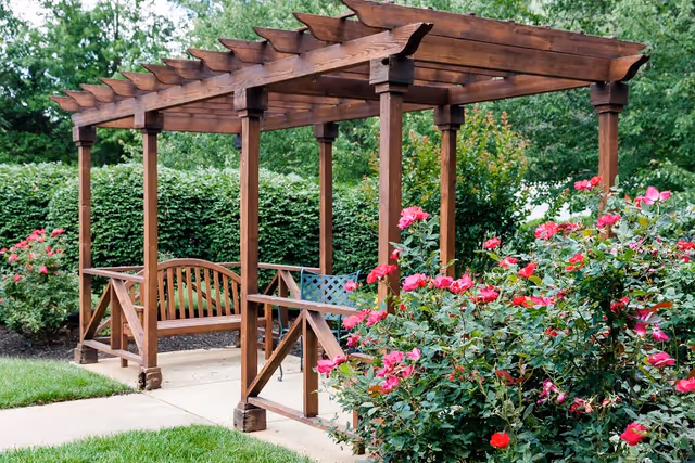 Wooden pergola with benches along a paved walkway, surrounded by pink rose bushes and greenery.