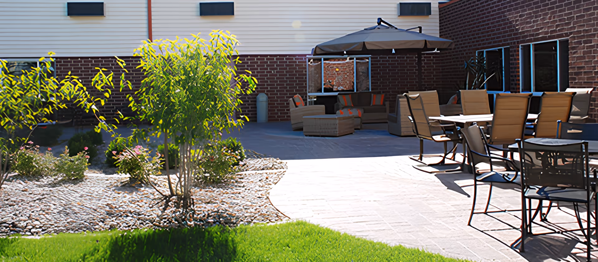 Sunny outdoor courtyard with patio dining chairs, lounge seating under an umbrella and landscaped planting beds beside a brick building.