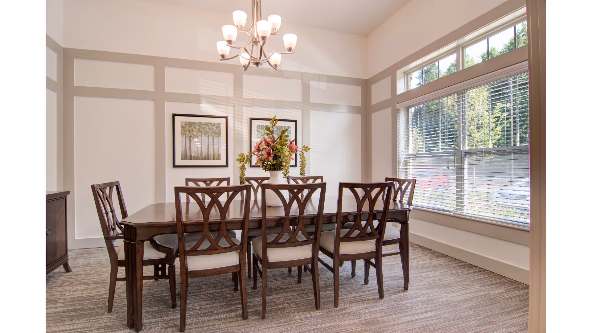 A bright dining room with a wooden dining table surrounded by six matching chairs with cushioned seats. A vase with pink and green flowers is centered on the table. The room has large windows with blinds letting in natural light, two framed pictures on the wall, and a modern chandelier hanging from the ceiling.