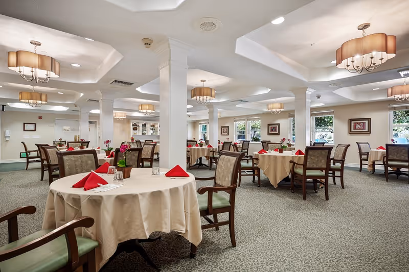 Spacious dining room with round tables covered in beige tablecloths, red napkins, wooden chairs, and chandeliers.