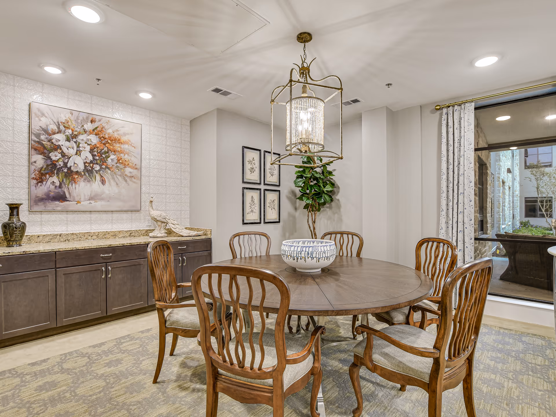 Round wooden dining table with six chairs under a crystal chandelier in a furnished dining room with artwork and a sideboard.