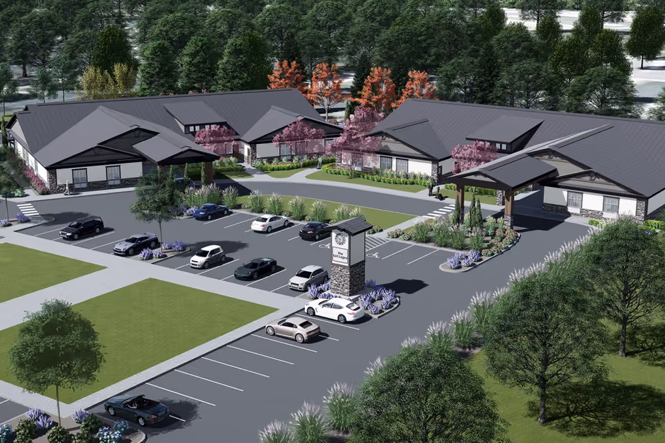 Aerial view of The Cottages Senior Care-Lochsa Falls, Meridian facility showing a single-story building with a dark gray roof surrounded by landscaped greenery and trees. The parking lot in front has several parked cars and a stone clock tower with the facility name. Walkways connect the building entrances to the parking area.