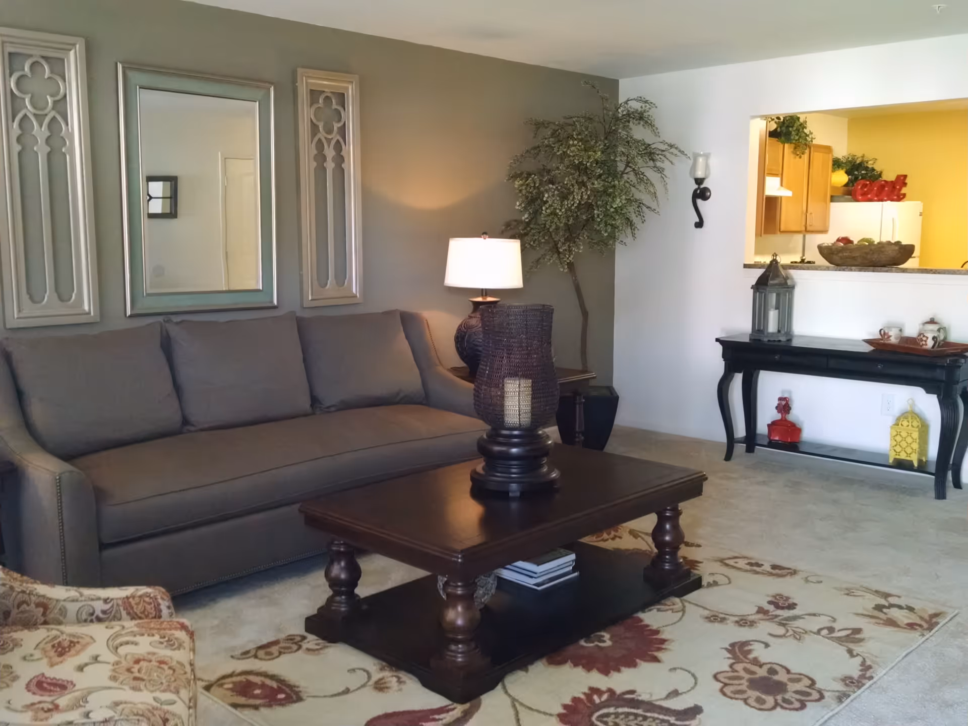A cozy living room with a gray sofa, a wooden coffee table with a decorative candle holder, a patterned armchair, a floor lamp, and a large potted plant. The room has a beige carpet with a floral rug, and a black console table against the wall with decorative items. A pass-through window reveals a kitchen area with wooden cabinets and a yellow wall with the word 'eat' displayed.