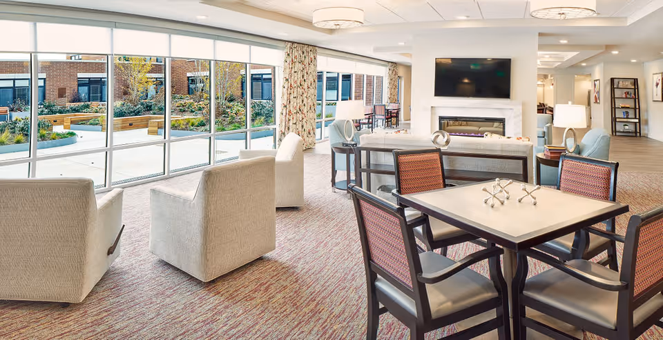 A bright and spacious living room area with large floor-to-ceiling windows overlooking an outdoor garden. The room features several beige armchairs, a table with four chairs, a fireplace with a mounted flat-screen TV above it, and modern decor including lamps and a bookshelf.