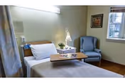 Furnished patient room with a single bed, overbed table, bedside lamp, armchair, and a window.