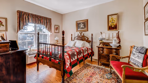 A cozy bedroom with a wooden bed frame featuring a red, green, and white patchwork quilt. The room has a large window with patterned curtains, a wooden dresser with framed pictures, a vintage nightstand with a lamp and decorative items, a red cushioned chair with a pillow, and a colorful patterned rug on the wooden floor.
