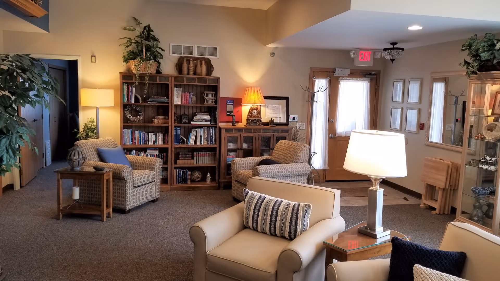 A cozy living room area in Timberwood Lodge Assisted Living featuring comfortable armchairs and a sofa with cushions, wooden bookshelves filled with books and decorative items, a side table with a lamp, indoor plants, and a glass display cabinet. The room has carpeted flooring, warm lighting, and a wooden door with a window covered by a sheer curtain.