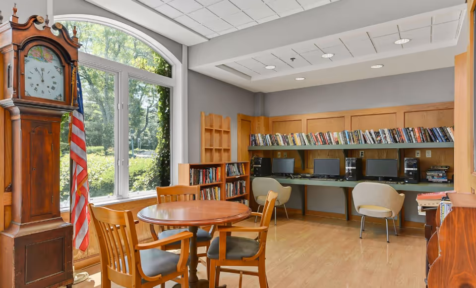 A cozy room with a round wooden table and four chairs in the foreground, a tall grandfather clock and an American flag near a large window showing greenery outside. In the background, there are bookshelves filled with books and a row of computers with chairs for use.