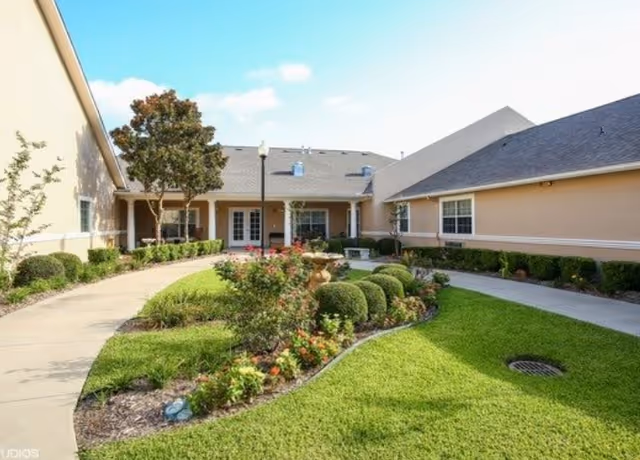 A landscaped outdoor courtyard area at Dogwood Trails Assisted Living and Memory Care featuring a curved walkway, neatly trimmed bushes, flowering plants, a small tree, and a central fountain. The courtyard is surrounded by beige buildings with windows and a clear blue sky overhead.