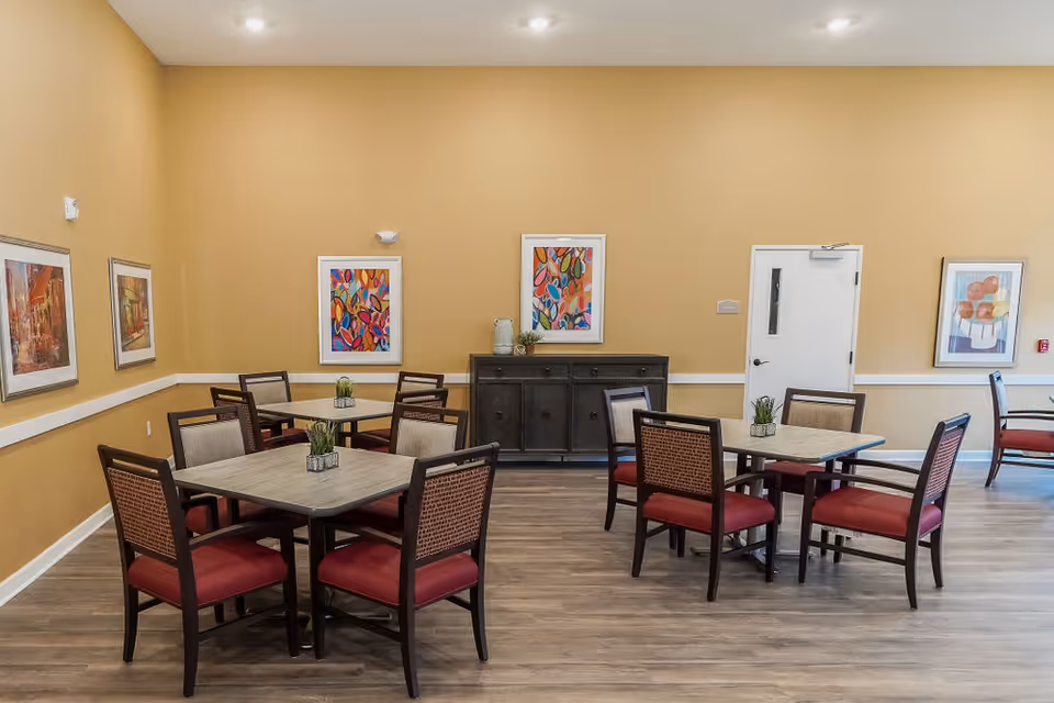 A dining room in a senior living facility with several square tables, each surrounded by four chairs with red cushions and woven backs. The walls are painted a warm beige color and decorated with colorful framed artwork. The floor is wood-style laminate, and there is a dark wooden sideboard against the far wall. The room is well-lit with recessed ceiling lights.
