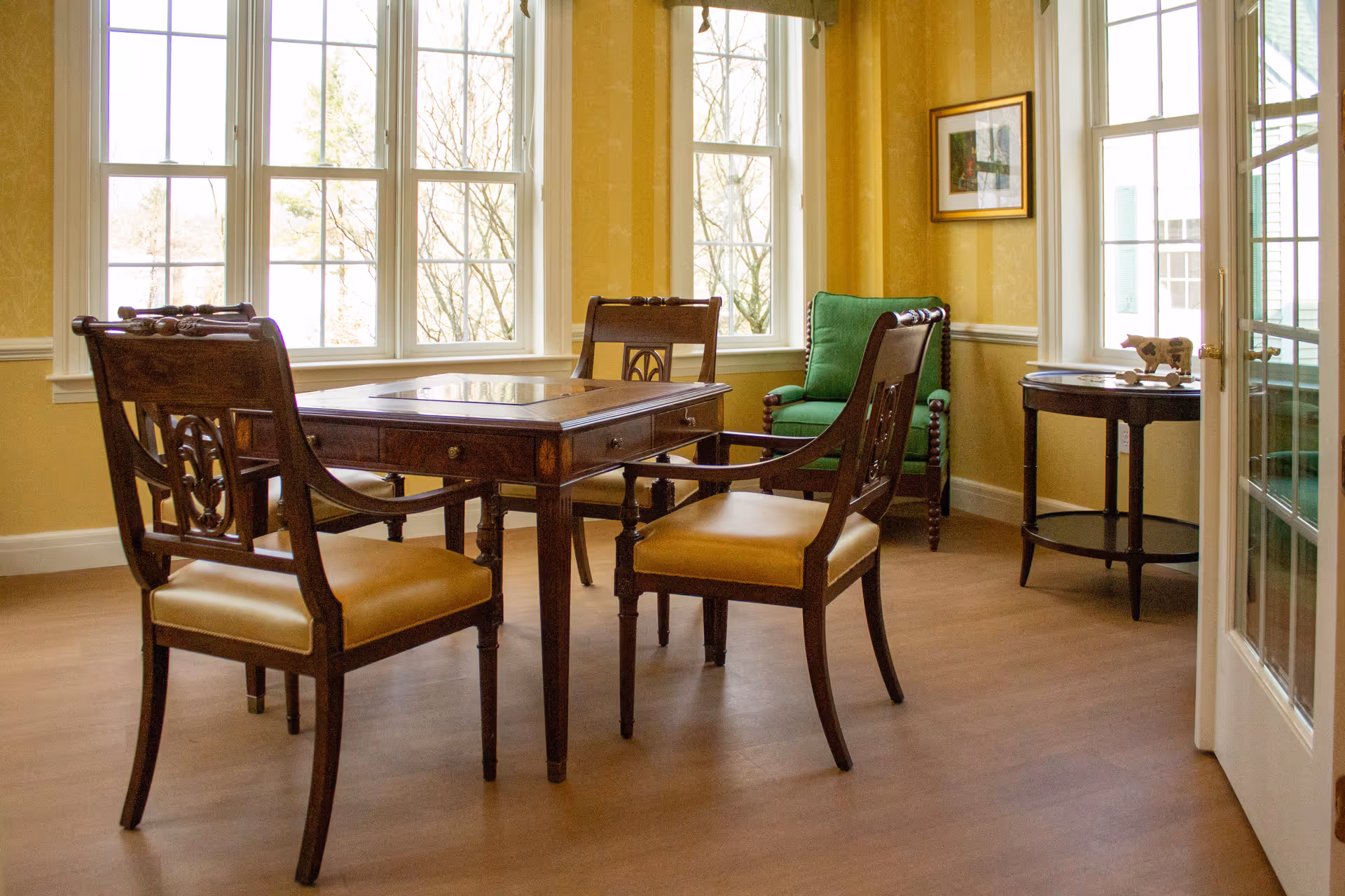 A bright room with large windows letting in natural light, featuring a wooden table with four matching wooden chairs with cushioned seats, a green upholstered armchair in the corner, and a small round side table with decorative items. The walls are painted yellow with white trim.