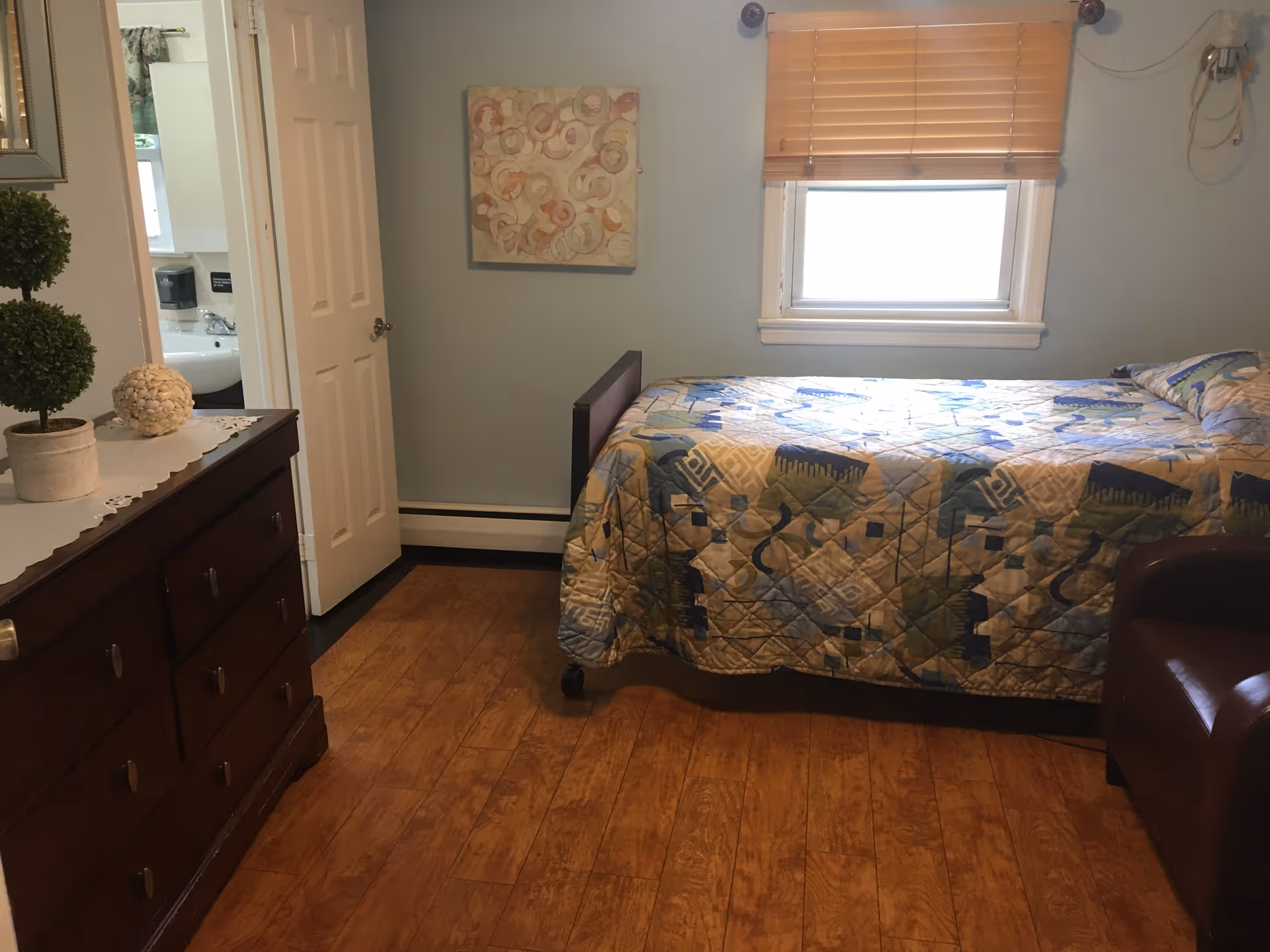 A small bedroom with a single bed covered in a patterned quilt, a window with wooden blinds, a brown armchair, and a wooden dresser with decorative items on top. A door is open showing a glimpse of a bathroom sink and mirror.