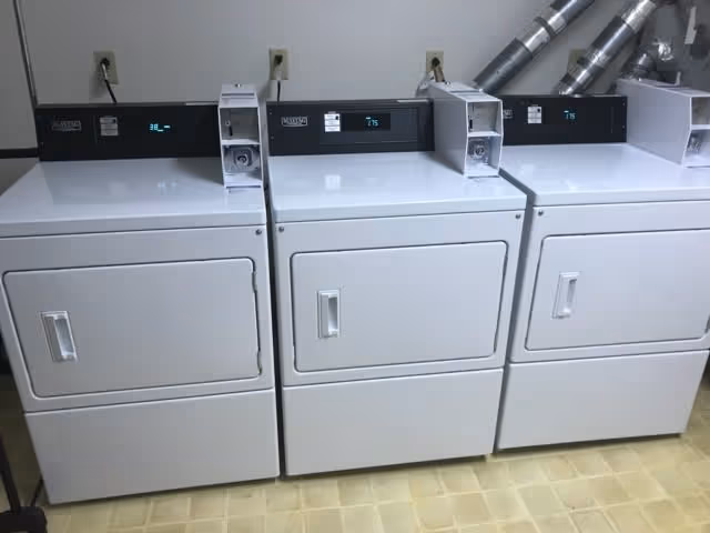 Three white coin-operated dryers lined up against a wall in a laundry room.