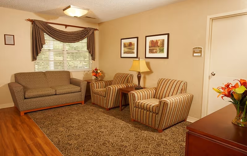 Cozy seating area with a sofa, two striped armchairs, side tables and a lamp in a carpeted senior living common room.