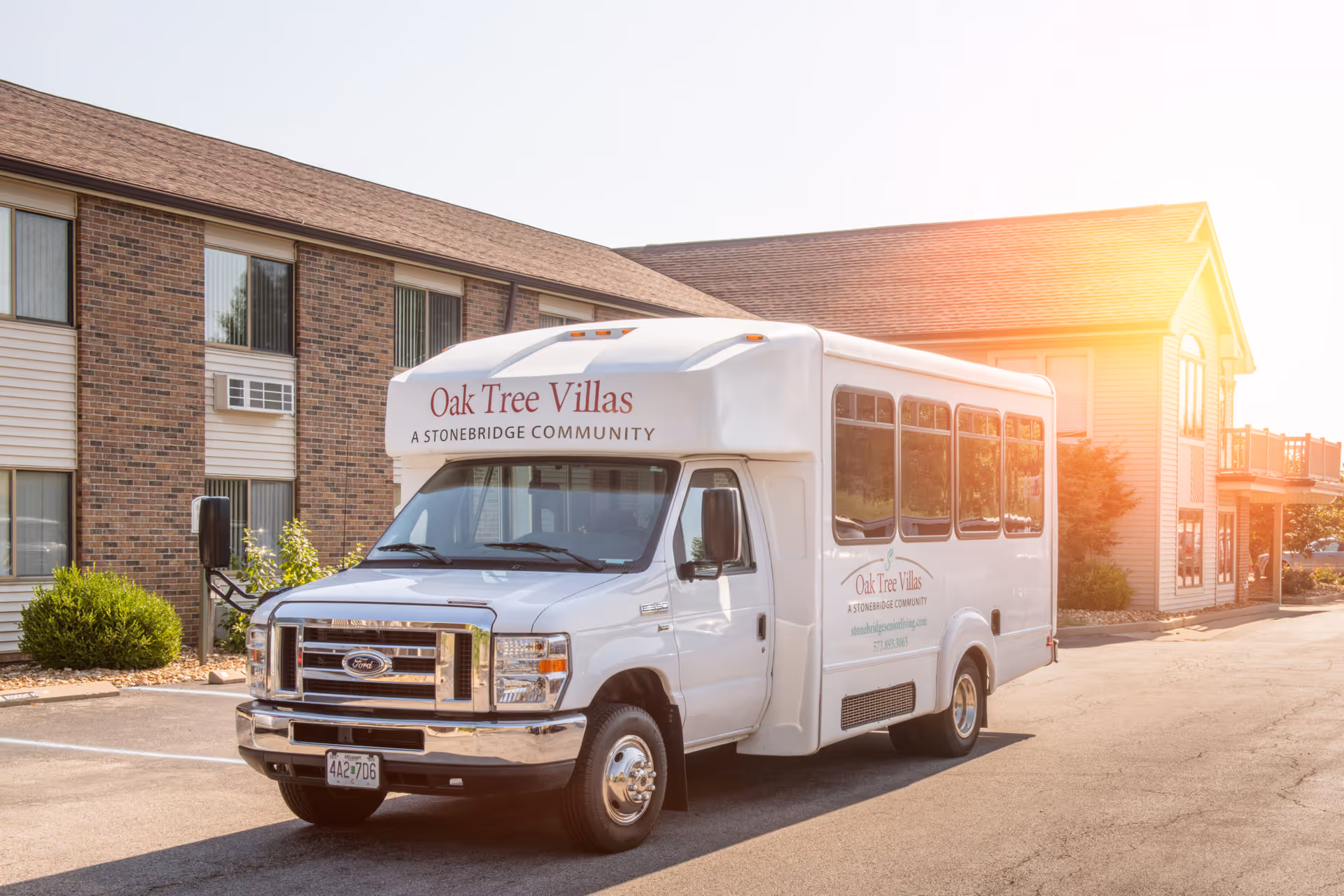 A white shuttle van labeled "Oak Tree Villas, A Stonebridge Community" parked outside a senior living building at sunset.