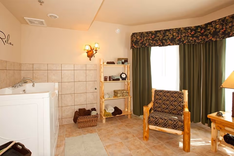 Bright senior living bathing room with a walk-in tub, tiled walls, wooden shelving and a rustic armchair by curtained windows.