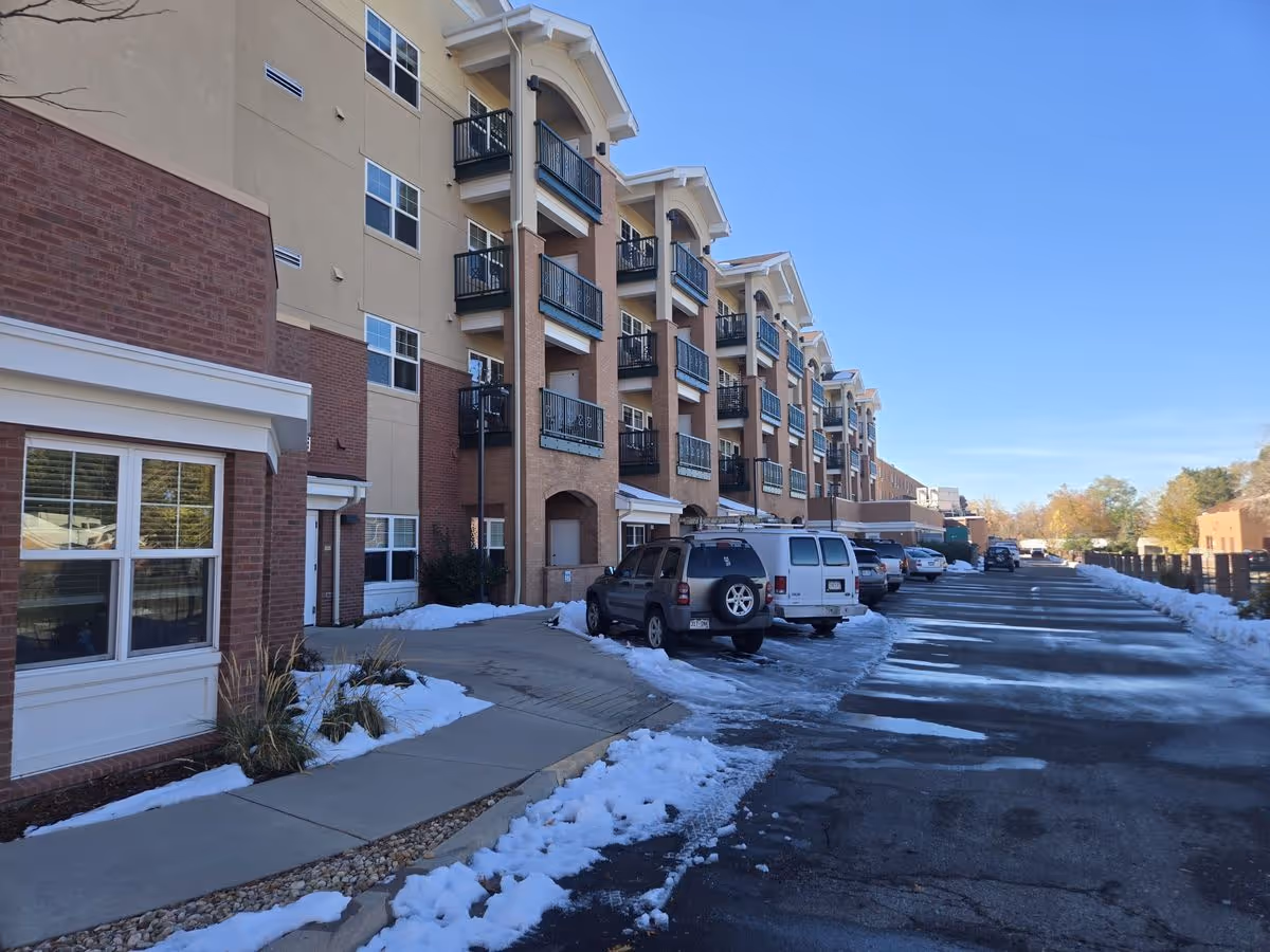 Front exterior of a multi-story residential care building with balconies, a snow-lined parking lot and parked cars.