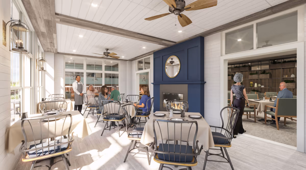 A bright dining room in a senior living facility with several tables covered in beige tablecloths and set with plates and glasses. There are black metal chairs with blue cushions around the tables. A blue fireplace with a round mirror above it is on one wall. Several seniors are seated and conversing, and a staff member in a white shirt and gray apron stands nearby. Another room with additional seating and plants is visible through a large opening. The room has white walls, wooden beams on the ceiling, ceiling fans, and large windows letting in natural light.