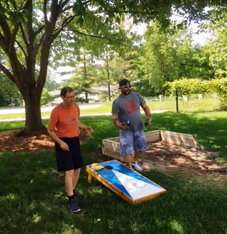 Two men playing cornhole outdoors on a grassy area with trees and a wooden cornhole board. One man is wearing an orange shirt and black shorts, and the other man is wearing a gray shirt, blue patterned shorts, and a black cap. The setting appears to be a sunny day in a garden or park-like area.