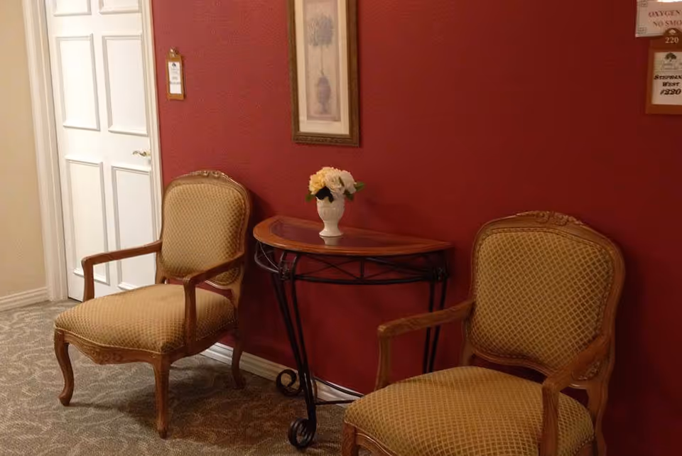 Two upholstered armchairs flank a small decorative console table with a vase of flowers against a red wall in an assisted living hallway.