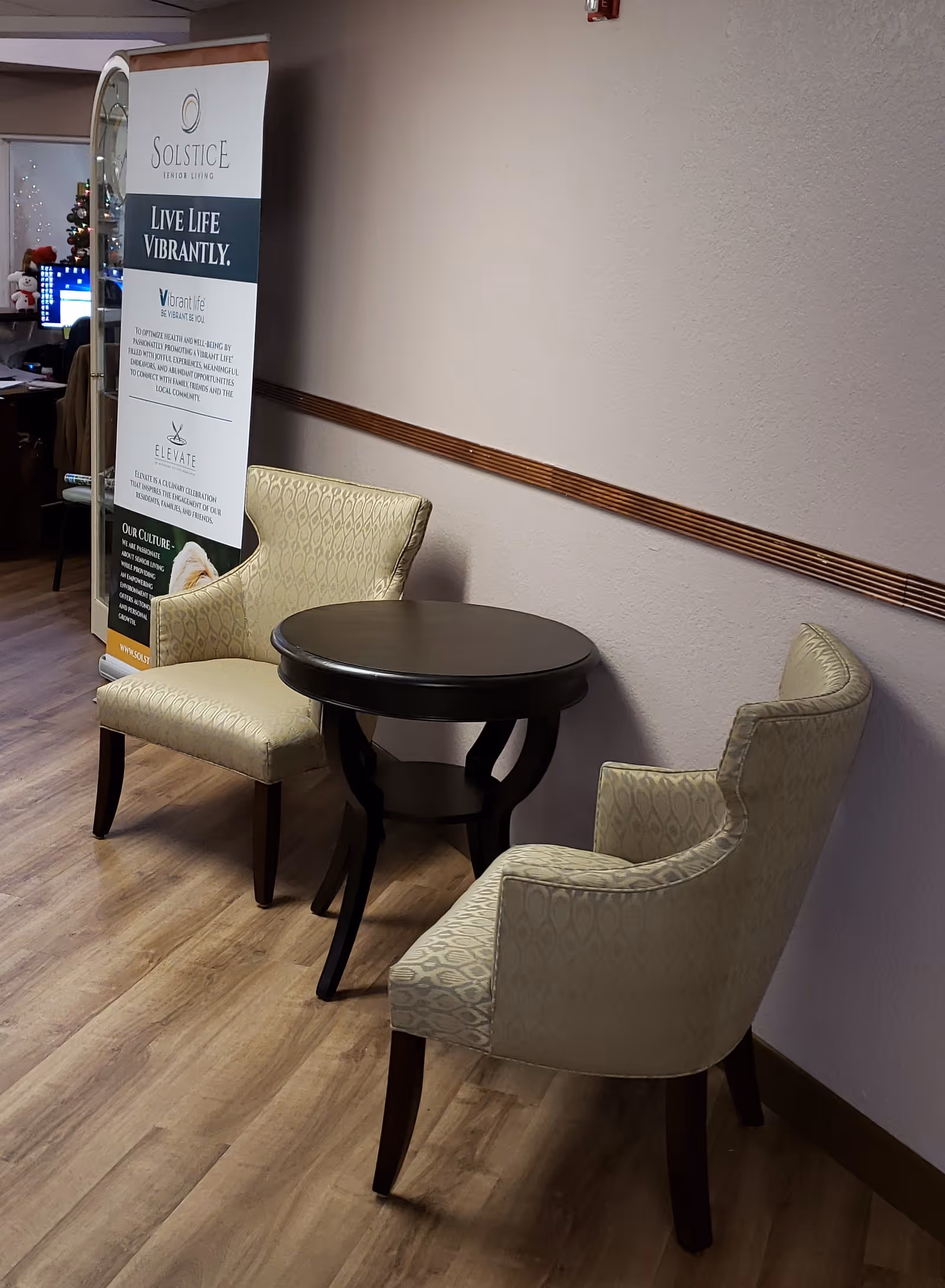 Two beige patterned armchairs positioned around a small round dark wooden table in a room with light-colored walls and wood flooring. A standing banner with the Solstice Senior Living logo and text is visible in the background near a doorway.