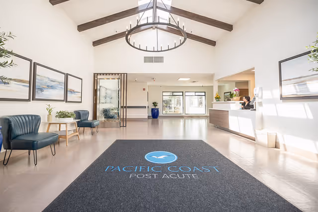 Bright and spacious reception area of Pacific Coast Post Acute facility with high vaulted ceiling and wooden beams. The floor features a large black mat with the facility's logo and name. There are blue chairs and small tables with plants along the left wall, framed artwork on the walls, and a reception desk on the right with a staff member seated behind it. Large windows allow natural light to fill the space.