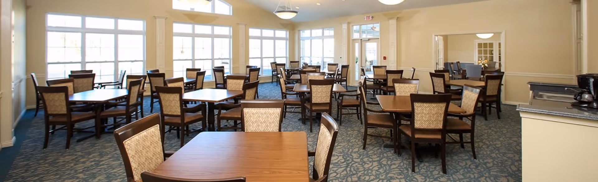 A spacious dining room with multiple wooden tables and chairs arranged neatly. Large windows allow natural light to fill the room, and there are light fixtures hanging from the ceiling. The room has a carpeted floor and light-colored walls.
