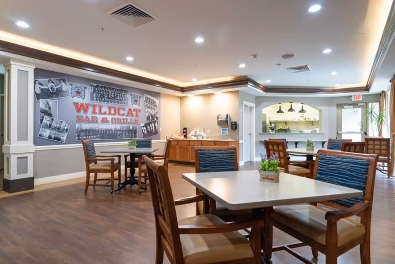 A dining area with several wooden tables and chairs with blue cushions. The walls are light-colored with decorative molding, and there is a large mural on one wall that reads 'WILDCAT BAR & GRILLE' surrounded by black and white photos. The ceiling has recessed lighting and crown molding with indirect lighting. In the background, there is a counter area with coffee and supplies, and an open kitchen window with hanging lights. A door with an exit sign is visible on the right side.