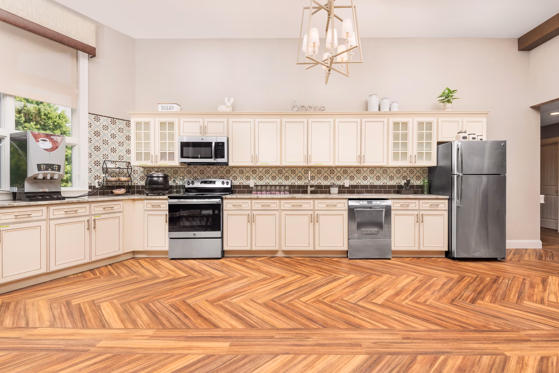 A spacious kitchen with cream-colored cabinets, stainless steel appliances including a refrigerator, oven, microwave, and dishwasher. The backsplash features a decorative tile pattern, and the floor has a wood herringbone design. A large window on the left lets in natural light, and a modern chandelier hangs from the ceiling.