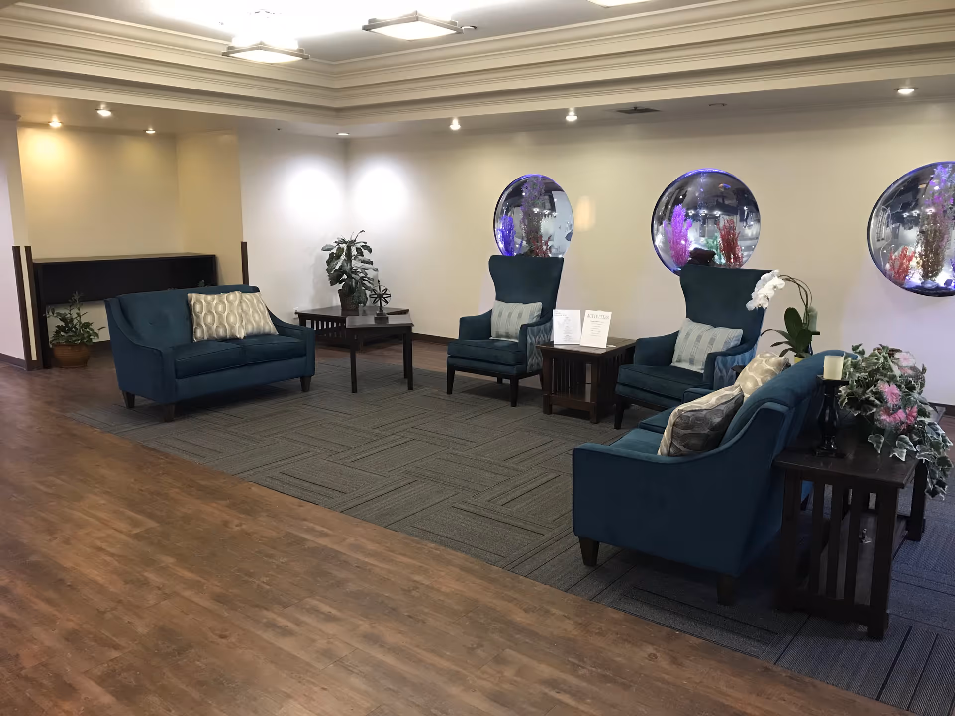 A seating area in a senior living facility with blue upholstered chairs and sofas arranged around wooden side tables. The room has a wooden floor and a carpeted section under the furniture. Three circular wall-mounted aquariums with colorful decorations are visible on the wall behind the chairs. There are also some potted plants and decorative items on the tables.
