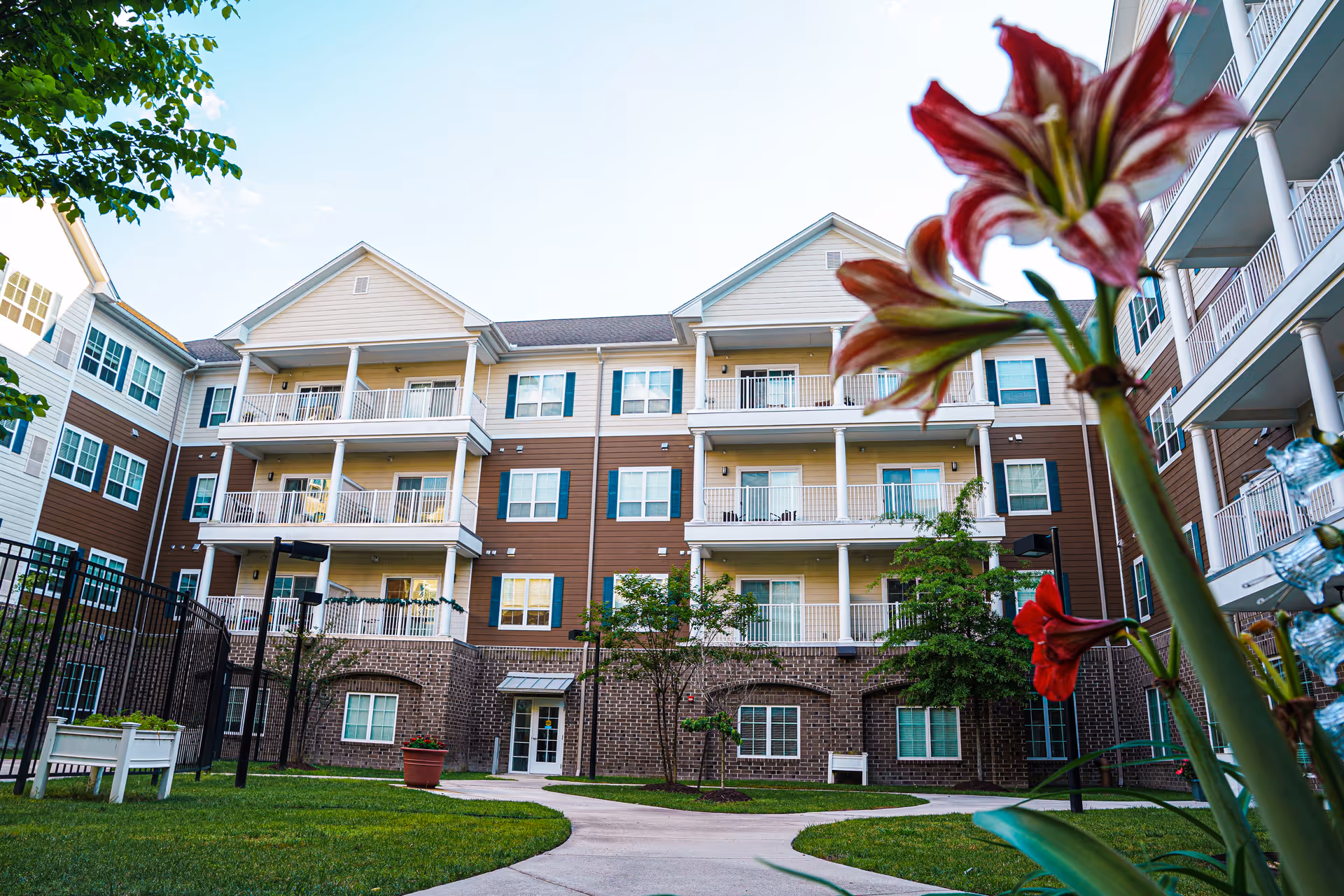 Exterior front view of a multi-story senior living building with balconies and a landscaped courtyard and flowers in the foreground.
