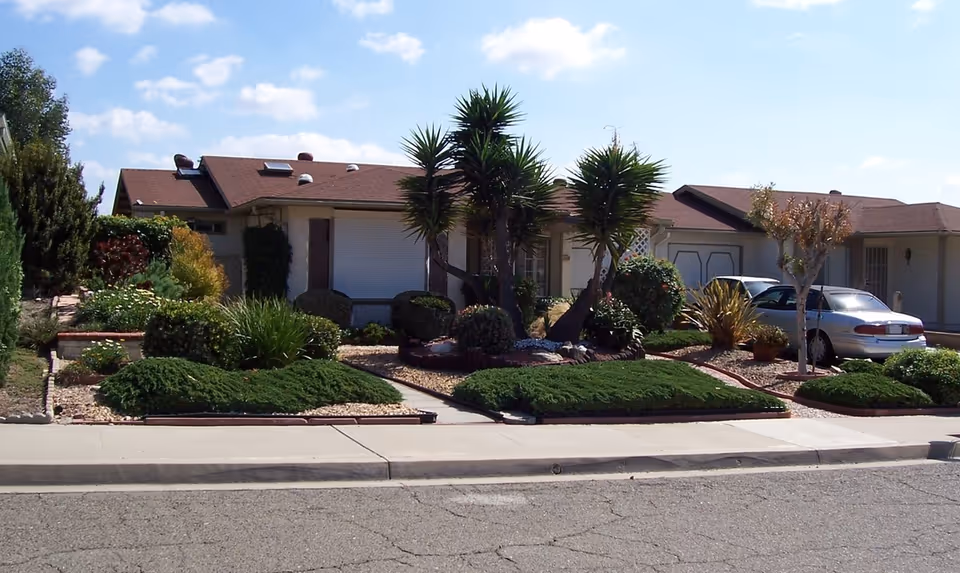 Front exterior of a single-story house with a landscaped yard, palm-like trees, and a car parked in the driveway.
