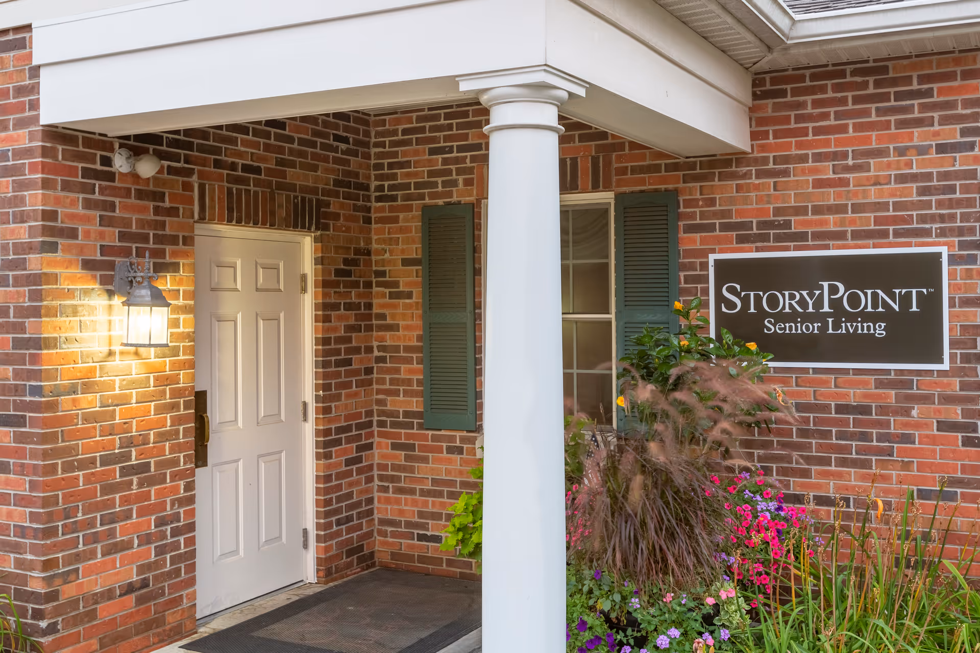 Entrance of StoryPoint Senior Living showing a white door, column, brick facade, sign, and flowering plants.