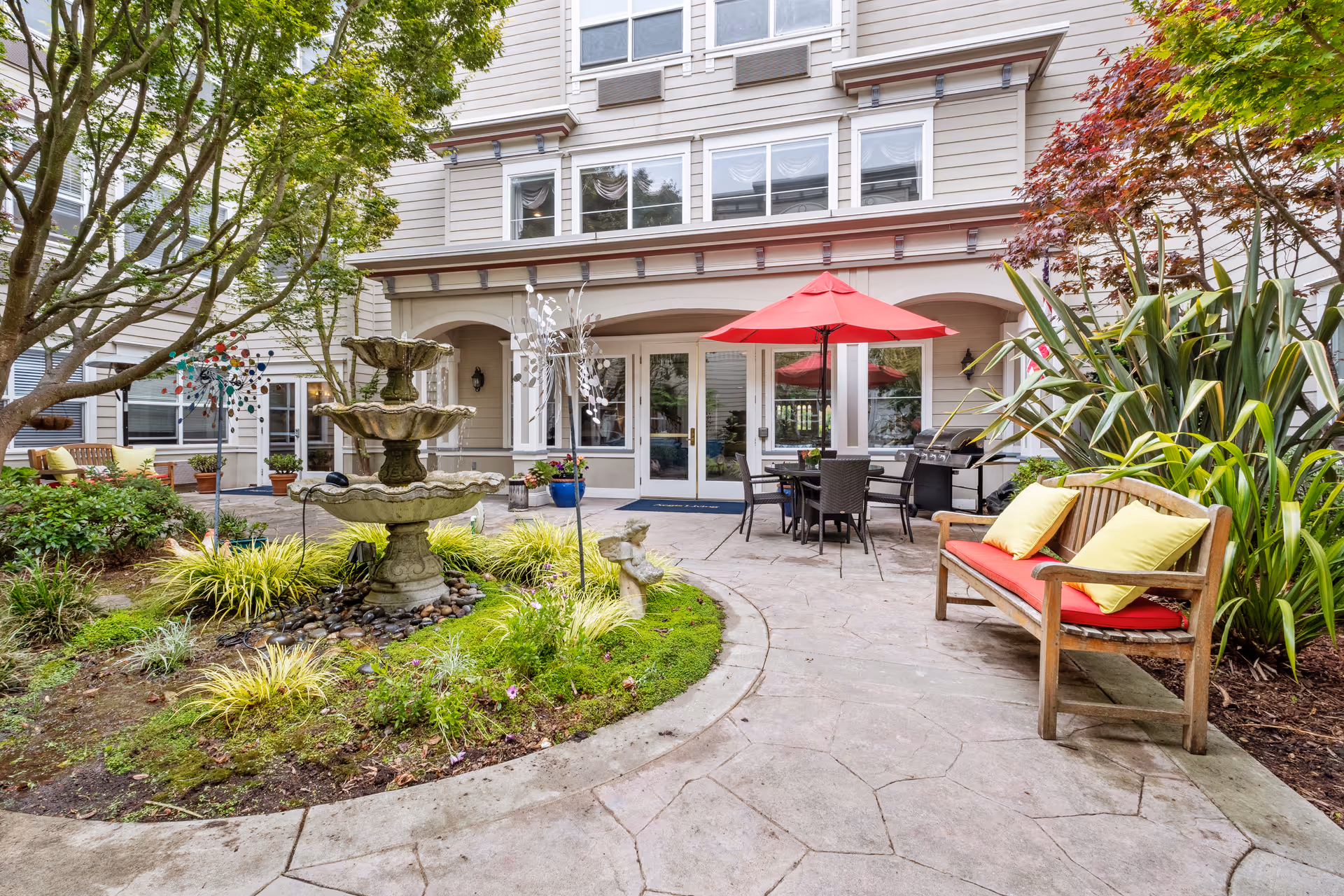 Outdoor patio area at Aegis Living San Francisco featuring a three-tier stone fountain surrounded by greenery, a wooden bench with red and yellow cushions, a table with chairs under a red umbrella, and a barbecue grill near the building entrance.