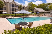 Outdoor swimming pool with patio tables, umbrellas, and lounge chairs in front of a multi-story residential building.