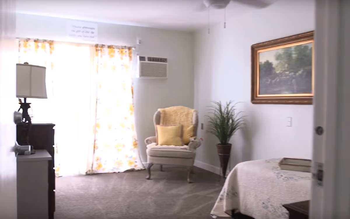 A cozy bedroom with a beige armchair adorned with yellow pillows and a throw blanket, a potted plant in a tall vase, a bed with a light-colored quilt, a dresser with a lamp, floral curtains covering a window, and a framed landscape painting on the wall.