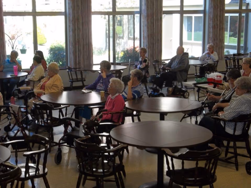 Several elderly residents seated at round tables in a bright common room with large windows.