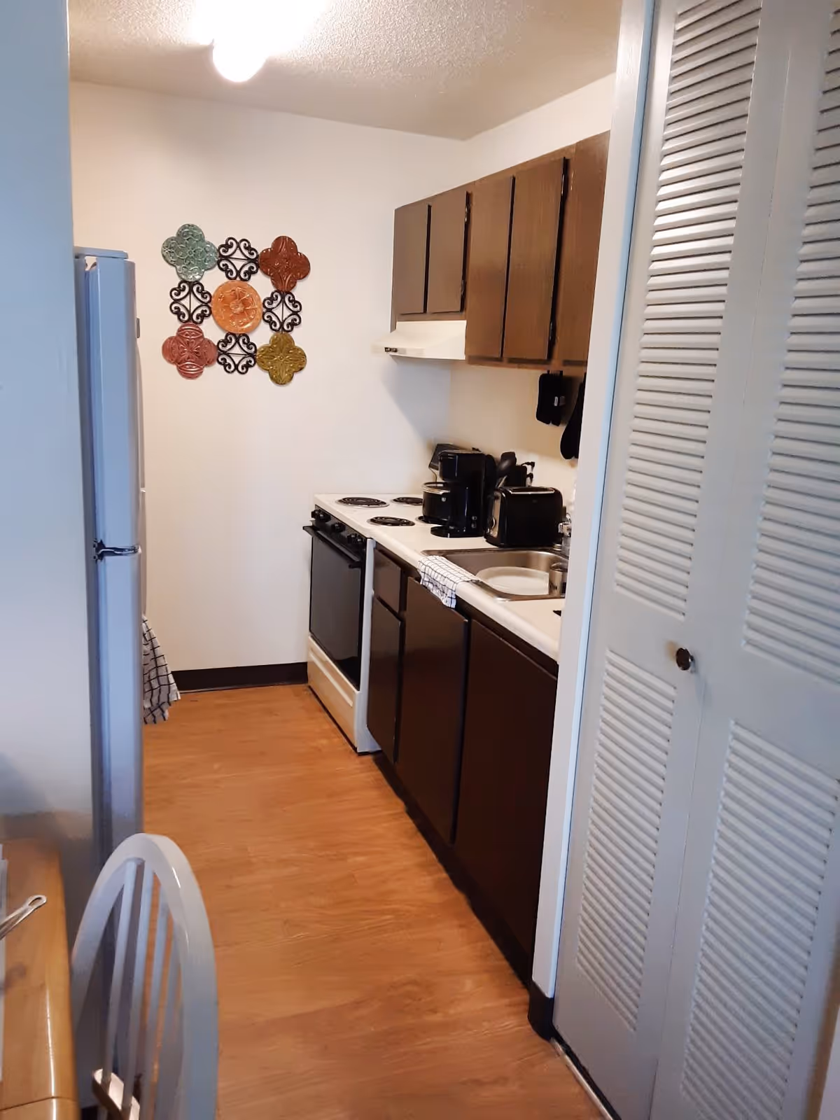Narrow galley kitchen with dark cabinets, stove, sink, refrigerator and decorative wall art on the far wall.