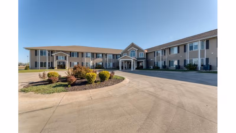Front exterior of a two-story beige retirement community building with a circular driveway and landscaped island under a clear blue sky.
