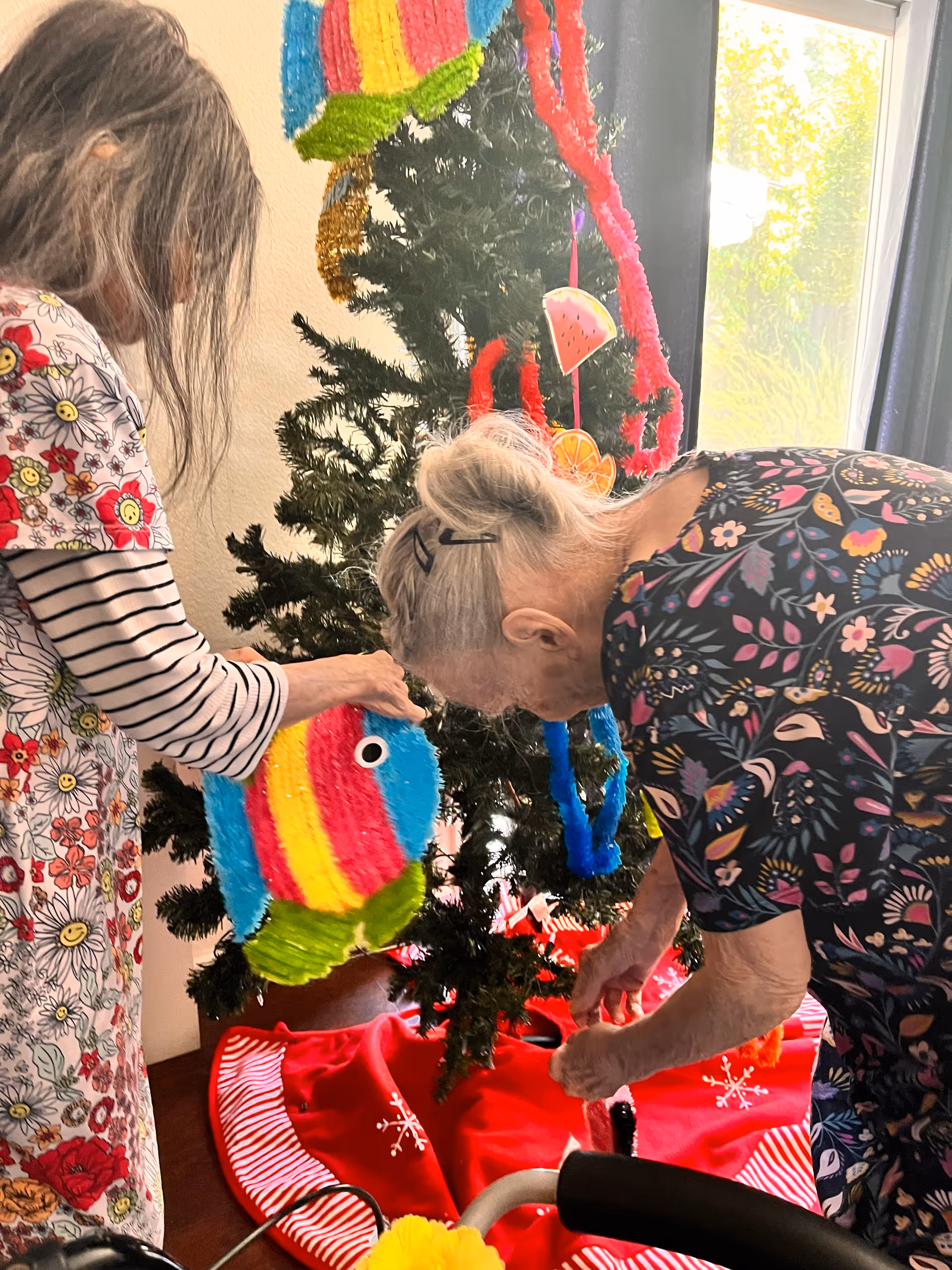 Two elderly women decorating a Christmas tree indoors. One woman with gray hair tied back is wearing a dark floral dress and is placing an ornament on the tree. The other woman, with long gray hair, is wearing a colorful floral and striped shirt and is holding a bright, multicolored fish-shaped decoration. The tree is adorned with colorful ornaments and garlands, and there is a red tree skirt with white snowflake patterns at the base. A window with natural light is visible in the background.